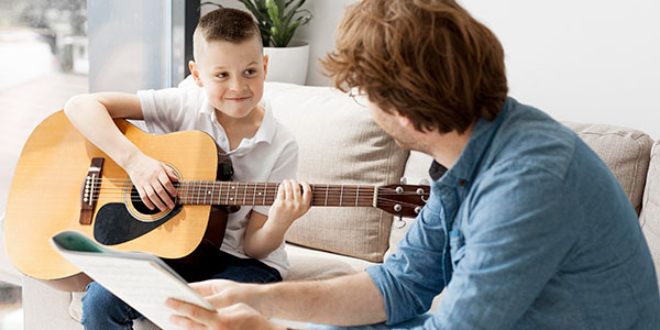 boy on couch playing guitar, man teacher sitting next to him pointing to music
